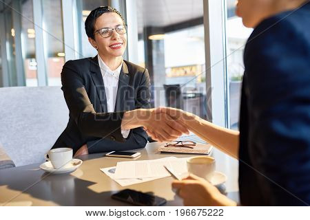 Portrait of confident female leader smiling happily while shaking hand of unrecognizable business partner during work meeting in cafe