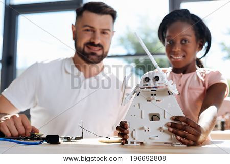 Best result. Adorable pre-teen girl holding a new white robotic warrior and smiling at the camera together with her robotics teacher, both being happy about the final result