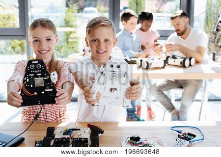 Gifted teens. Happy smiling siblings standing near the working table during their robotics class and holding their robot models of cosmic warriors while their teacher explaining something to others