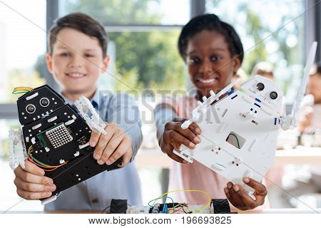 Bright smiles. Happy pre-teen boy and girl standing in the robotics classroom and posing with their robot models, having created them by themselves