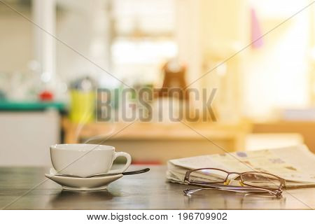 Closeup cup of hot cappuccino coffee with newspaper and spectacles on the wood table with blur kitchen room background-warm tone.