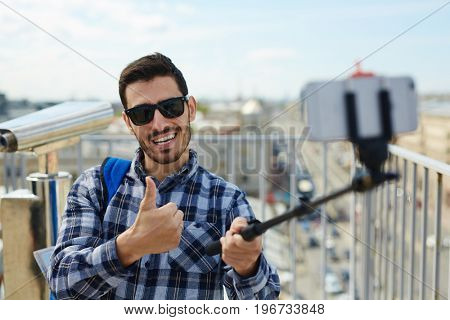 Portrait of handsome young man taking selfie photo while standing on rooftop against panoramic city view and coin-operated binoculars in background
