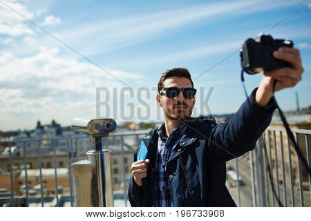 Portrait of handsome young man taking selfie photo while standing on rooftop viewing platform against panoramic city view and coin-operated binoculars in background