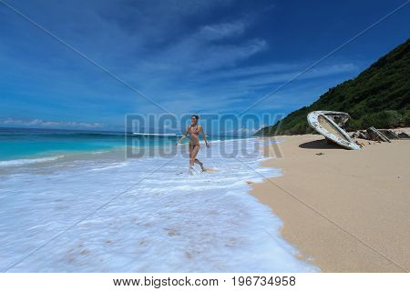 A young woman in bikini runs along an ocean beach with waves.On the beach lie the wreckage of a boat