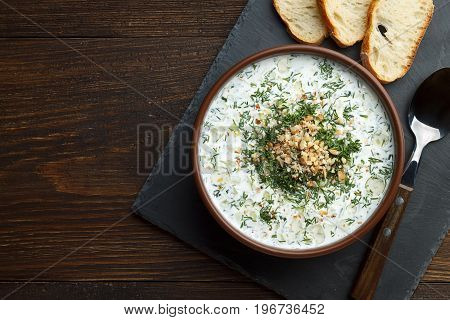 Traditional bulgarian chilled tarator in brown bowl with spoon and bread ready to eat. Summer cold soup with cucumber yogurt dill and walnuts on wooden table.