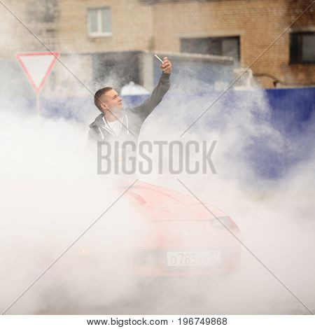 Orel Russia July 22 2017: Dynamica car festival. Young man making selfie in burning wheel smoke clouds with industrial background
