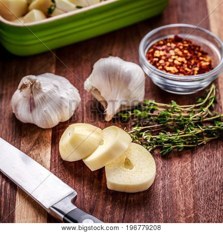 Preparing ingredients for roasted garlic includes fresh thyme and crushed red pepper. Chopped garlic in in a ceramic baking dish. Knife and roasting pan also shown