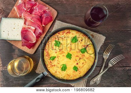 An overhead photo of a Spanish tortilla with potatoes and zucchinis in a tortillera, with jamon, cheese, glasses of white and red wine, and two forks, with a place for text