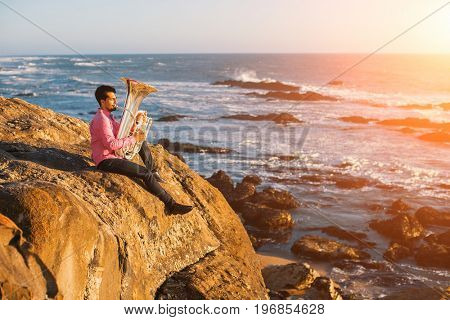Young musician play the trumpet on rocky sea coast during surf. Tuba instrument. 
