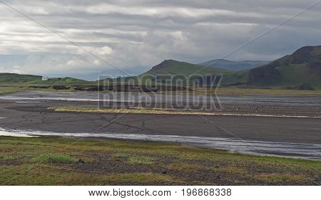 Isolated White House In The Wild Iceland Landskape With Green Hills, Grass, Puddle, Dirt And Puffy S