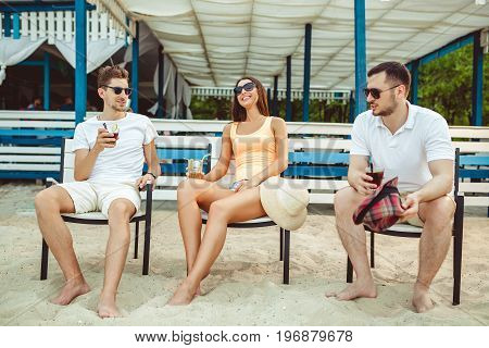 Lifestyle Young people enjoying summer vacation sunbathing drinking at beach bar. Two guys and a young girl sit on the terrace of the summer bar and enjoy the conversation