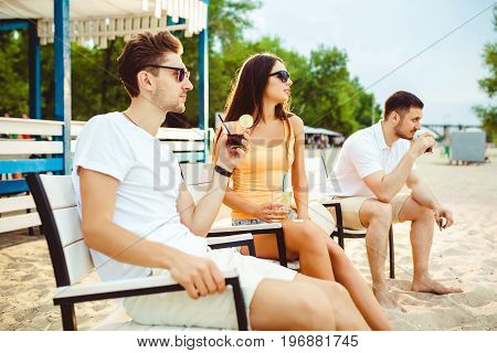 Lifestyle Young people enjoying summer vacation sunbathing drinking at beach bar. Two guys and a young girl sit on the terrace of the summer bar and enjoy the conversation