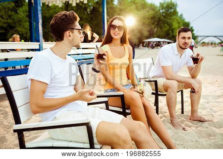 Lifestyle Young people enjoying summer vacation sunbathing drinking at beach bar. Two guys and a young girl sit on the terrace of the summer bar and enjoy the conversation