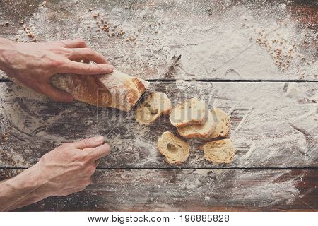 Baking background. Hands of baker cutting bread loaf with knife on rustic wooden table sprinkled with flour.