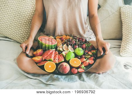 Summer healthy raw vegan clean eating breakfast in bed concept. Young girl wearing pastel colored home clothes sitting and holding tray full of fresh seasonal fruit