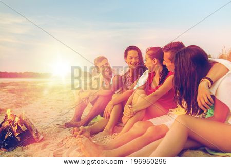 friendship, happiness, summer vacation, holidays and people concept - group of smiling friends sitting near fire on beach