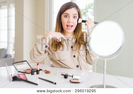 Beautiful young woman using make up cosmetics applying color using brush with surprise face pointing finger to himself