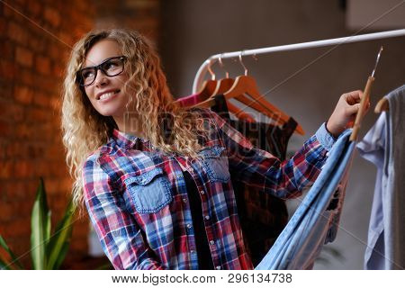 Smiling Cheerful Woman In Glasses And Checkered Shirt Chooses Clothes In Butique  For Her Holidays. 