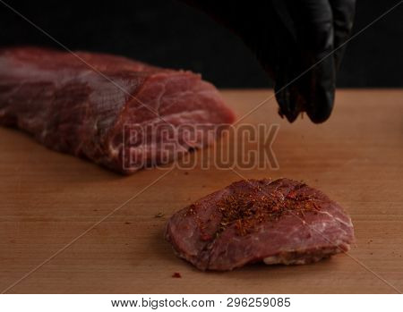Hand In Black Glove Sprinkling Spice On Beef Steak On A Wooden Board With A Black Background