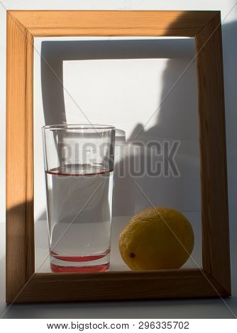 Glass Of Crystal Clear Water In The Light Of The Sun Next To A Lemon In A Wooden Frame On A White Ba