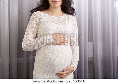 Image Of Pregnant Woman Standing In White Gentle Dress And Touching Her Belly Against Window With Cu