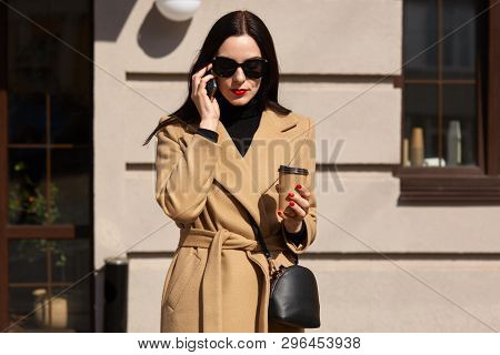 Outdoor Shot Of Serious Lovely Attractive Model Poses Over Local Restaurant, Holding Phone In Her Ha