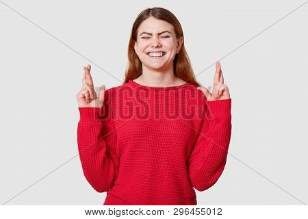 Indoor Shot Of Pleasant Looking Dressed In Casual Red Sweater, Crosses Fingers And Has Great Hope Fo