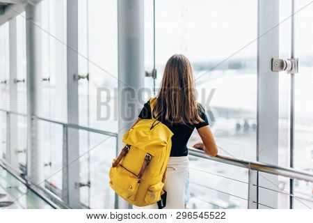 Teen girl looking at the window while waiting for international flight in airport departure terminal. Young passenger with backpack travelling on airplane. Teenager tourizm abroad alone concept.