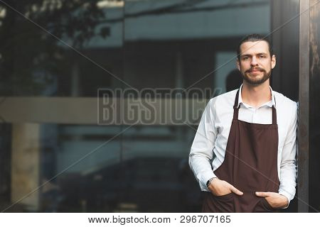 Successful Sme. Male Barista Leaning Against On Front Of The Coffee Shop Glass Wall. Portrait Of Bea
