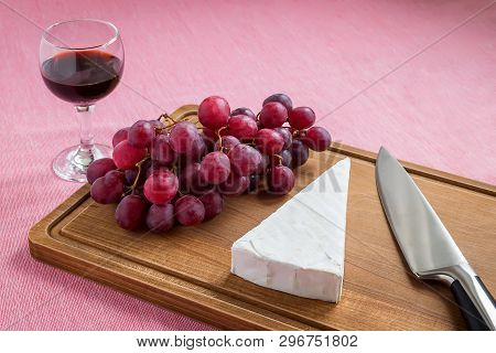 Triangular Piece Of Brie Cheese, Red Sweet Grapes, Chef Knife On A Brown Wooden Cutting Board And A 