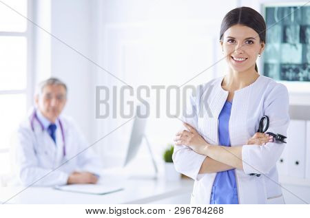 A doctor smiling at the camera with her male colleage in the back of the consulting room in hospital