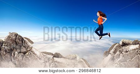 Redhead Woman Jumps Through The Gap Between Hills Over Cliff On Blue Sky Background.
