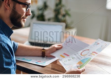 Serious young economist looking through financial papers with charts and diagrams while sitting by desk