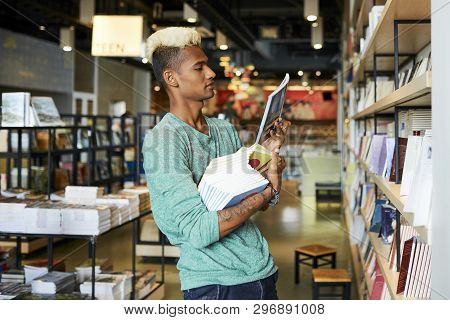 Serious Handsome Young Black Bookstore Manager With Blond Mohawk Holding Stack Of Books And Checking