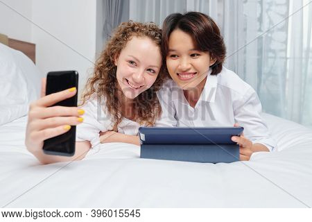 Joyful Multi-ethnic Female Friends Lying On Bed With Tablet Computer And Taking Selfie On Smartphone