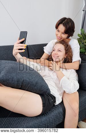 Happy Young Multi-ethnic Lesbian Couple Cuddling On Sofa And Taking Selfie
