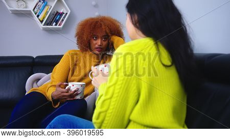 Two Young Female Friends With Coffee Conversing In The Living Room. African American And Asian Women