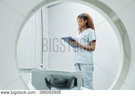 Young African nurse in uniform making notes in medical document while standing by couchette of ultra sound equipment before new patient