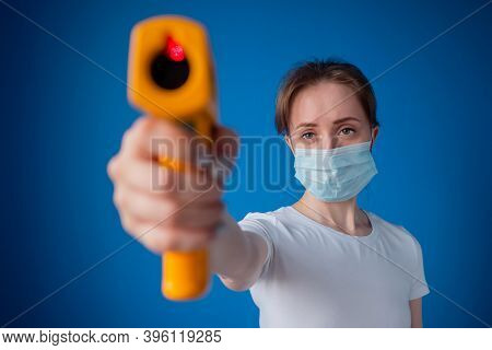 Woman In Medical Face Mask Holding Yellow Pyrometer To Measure Temperature Toward Camera - Close Up 