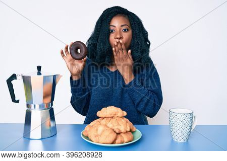 Beautiful african woman eating breakfast holding cholate donut covering mouth with hand, shocked and afraid for mistake. surprised expression 