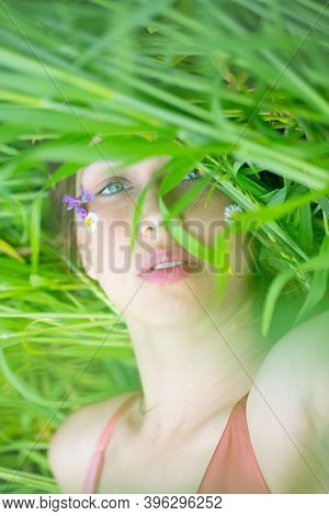 Beautiful Girl Lies On Green Wheat Field With Wild Flowers On Her Face. Search For Harmony, Healthy 