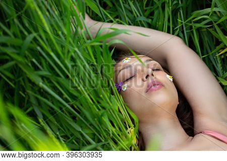 Beautiful Girl Lies Eyes Closed On Green Wheat Field With Wild Flowers On Her Face. Search For Harmo