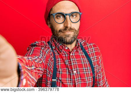 Handsome hipster man with beard taking a selfie photo with smartphone thinking attitude and sober expression looking self confident 