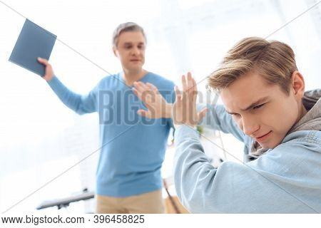Father Swings To Hit His Son With Book. Troubled Teenager Covers Himself With Hands.