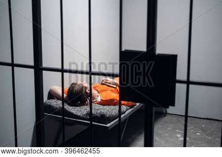 Lights Out In Prison. A Convict In An Orange Robe Rests On A Bunk In A Cell With A Book