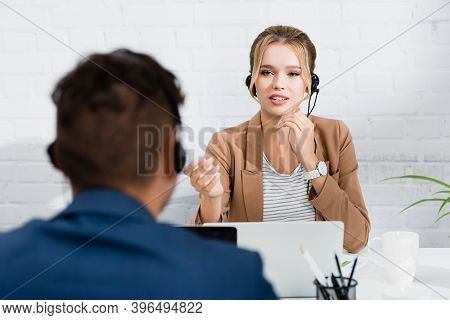 Consultant In Headset Gesturing, While Sitting At Workplace On Blurred Foreground