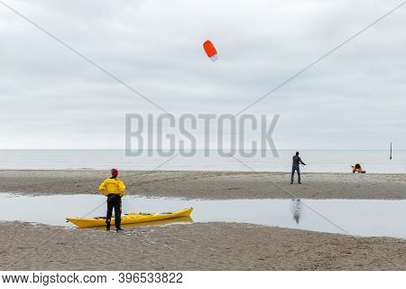 Koksijde, Belgium - November 8, 2020: A Man Standing Next To A Yellow Kayak, Watches Another Man Fly