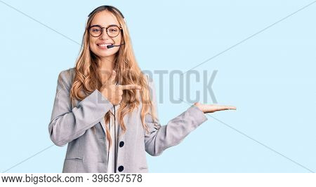 Young beautiful blonde woman wearing call center agent headset amazed and smiling to the camera while presenting with hand and pointing with finger. 