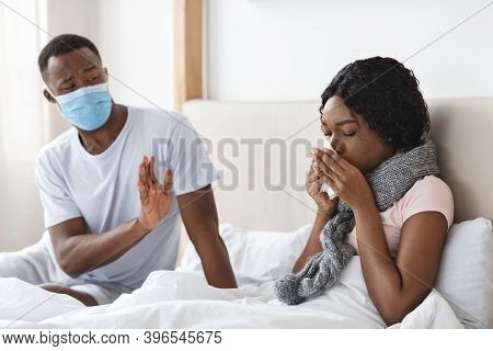 Black Man Wearing Protective Face Mask While His Wife Is Sick, Bedroom Interior, Empty Space. Africa