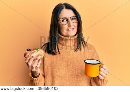 Middle age brunette woman eating pastry and drinking coffee relaxed with serious expression on face. simple and natural looking at the camera. 